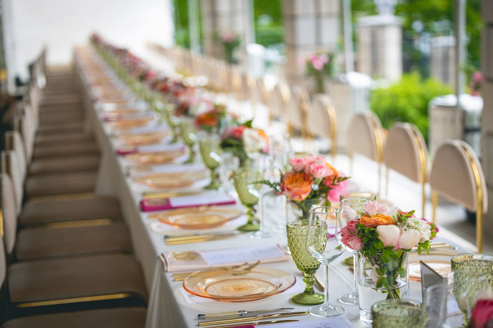 Longue table à dîner pour une grande liste d'invités, avec des assiettes transparentes colorées, des verres à pied verts accompagnés de verres à vin et de petits bouquets de fleurs dans les tons roses et oranges