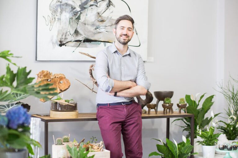 Homme souriant qui croise les bras et qui prend la pose assis contre un présentoir de fleuriste avec des éléments de décoration design, des plantes vertes, grasses et des fleurs